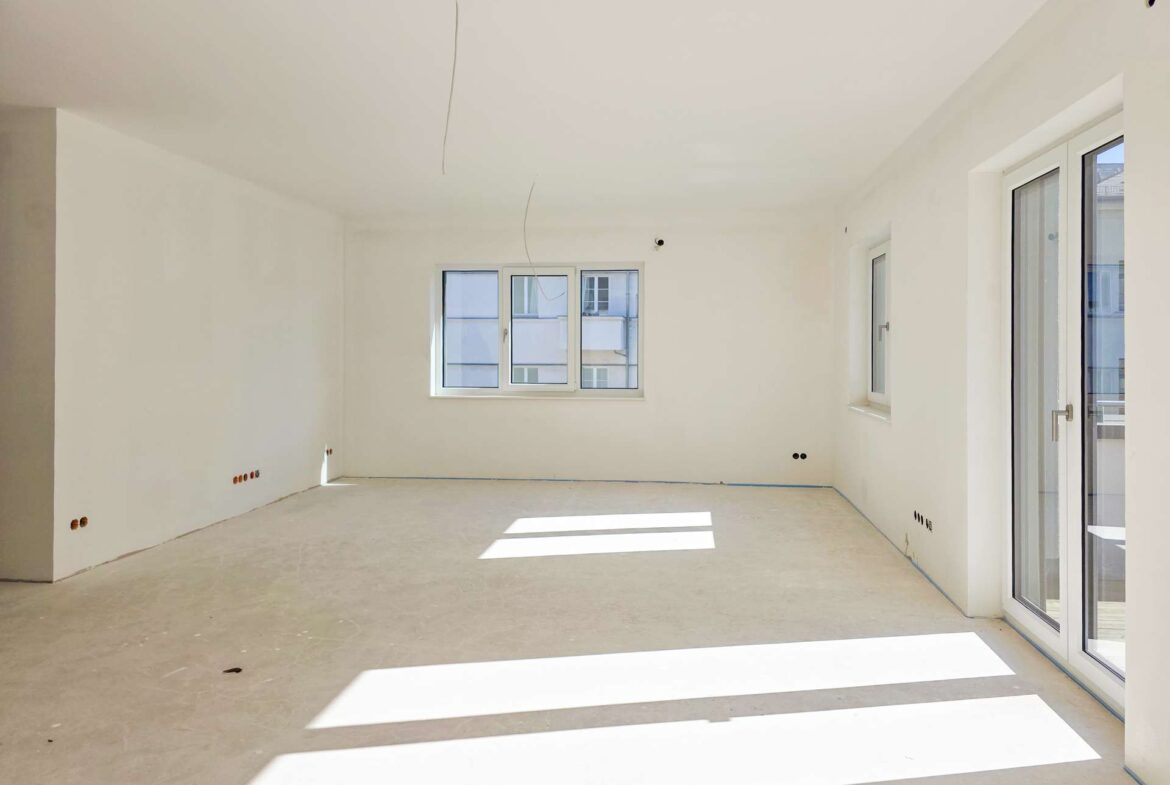 Empty, unfinished room with white walls, exposed ceiling wires, and large windows letting in sunlight