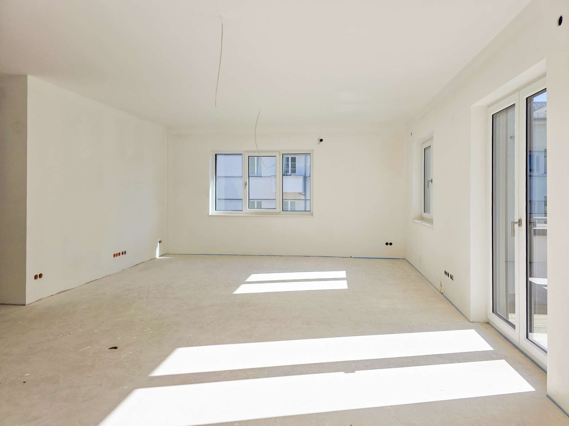 Empty, unfinished room with white walls, exposed ceiling wires, and large windows letting in sunlight