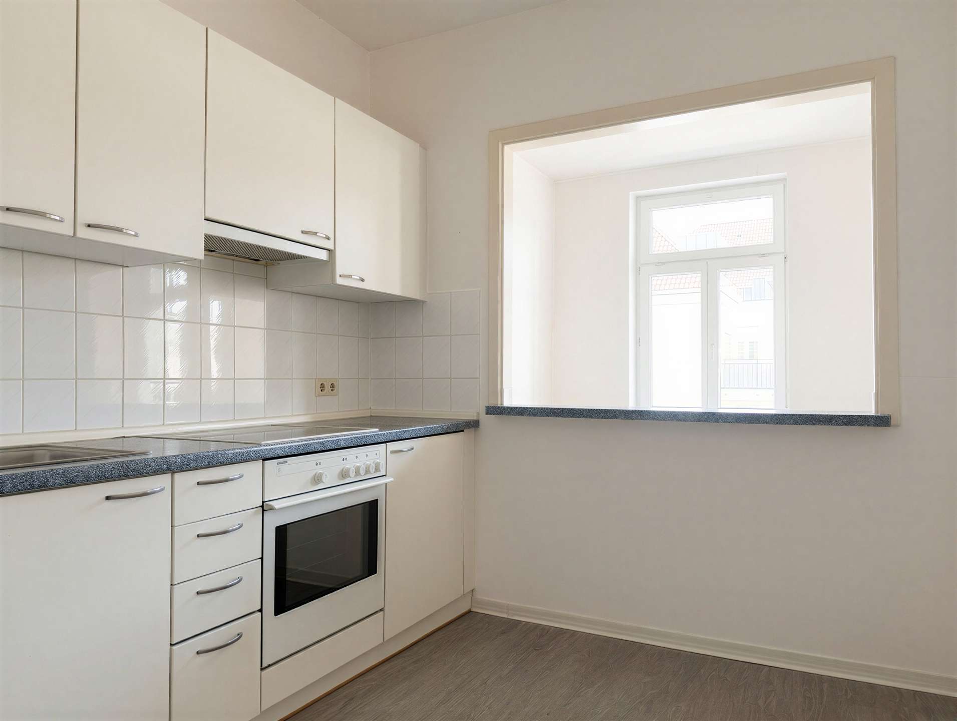 White kitchen with upper cabinets, a built-in oven, and a gray speckled counter; a pass-through window to a bright adjoining room.