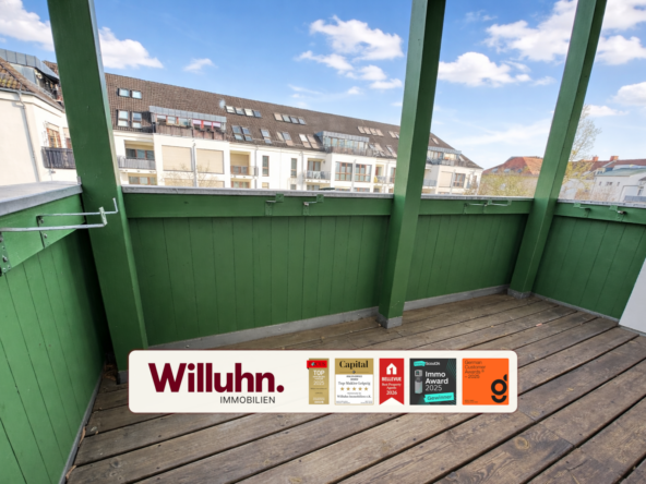 Green wooden balcony with a railing overlooking a residential street and blue sky; Willuhn Immobilien sign on the floorboards in the foreground.