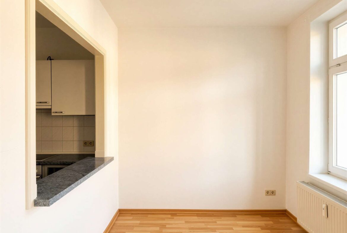 Empty sunlit room with wood flooring, a large window, and a kitchen pass-through with granite counter on the left.