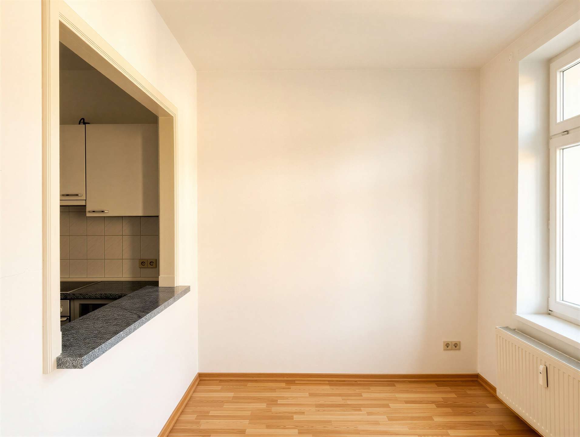 Empty sunlit room with wood flooring, a large window, and a kitchen pass-through with granite counter on the left.