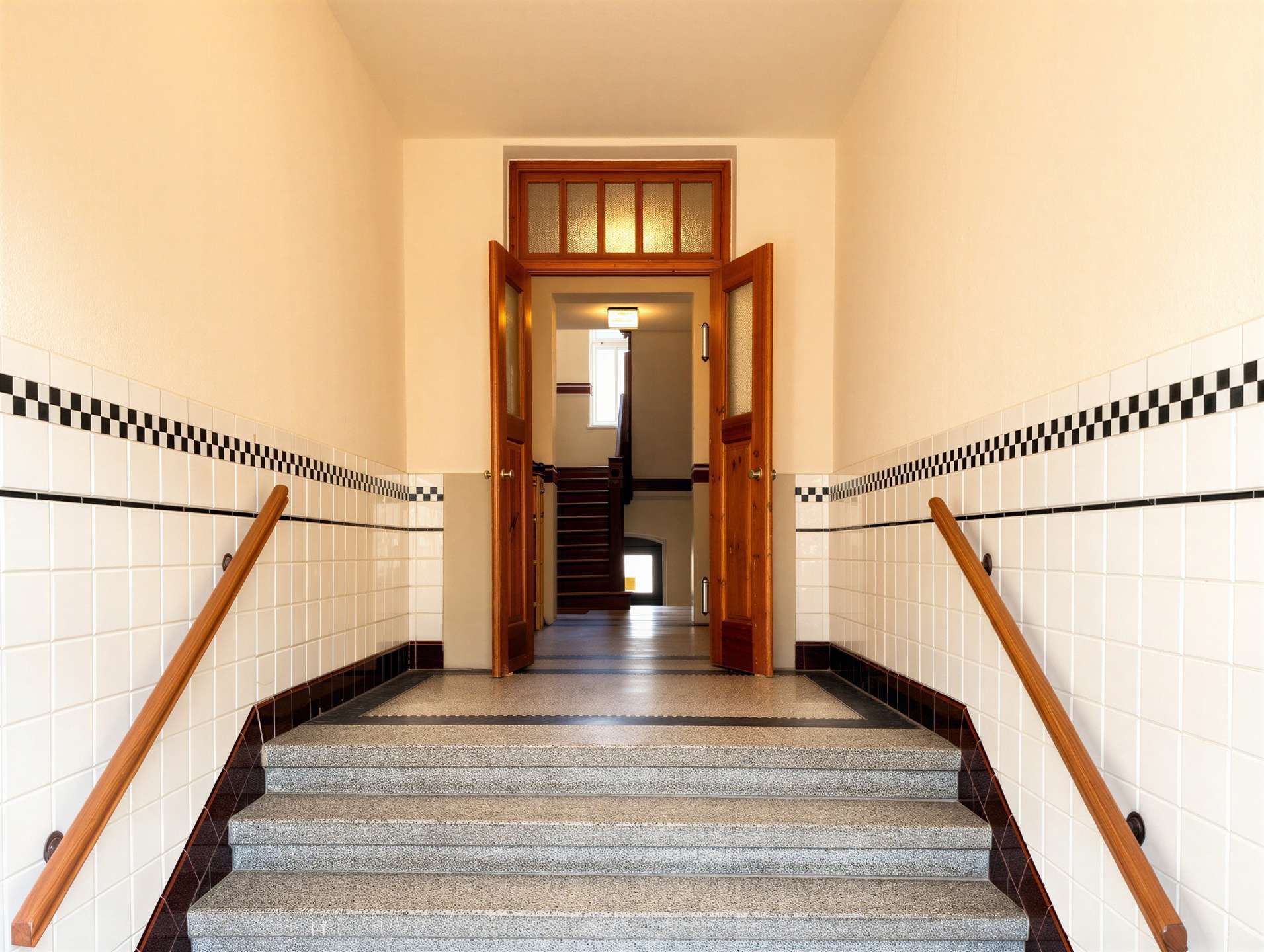 Indoor stairwell with tiled walls and checkered pattern, wooden handrails, and double doors opening to a lit hallway.