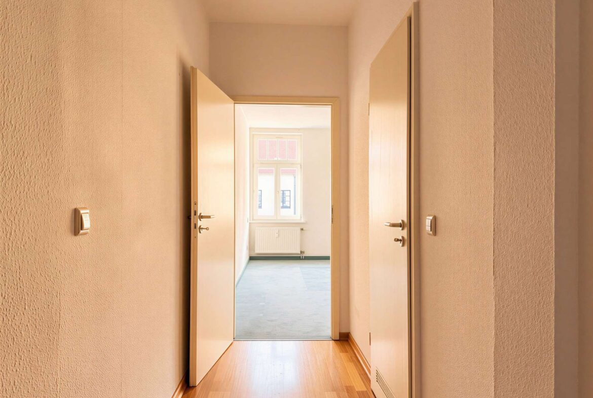 Narrow beige hallway with two open doors, opening to a sunlit room with a window and radiator at the far end