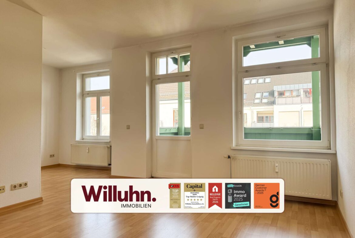 Bright empty room with large windows letting in sunlight, wooden floor, and a long Willuhn Immobilien sign with award stickers on the floor.