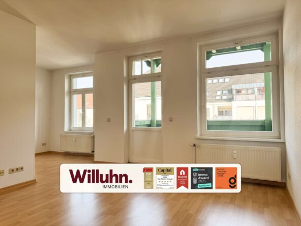 Bright empty room with large windows letting in sunlight, wooden floor, and a long Willuhn Immobilien sign with award stickers on the floor.