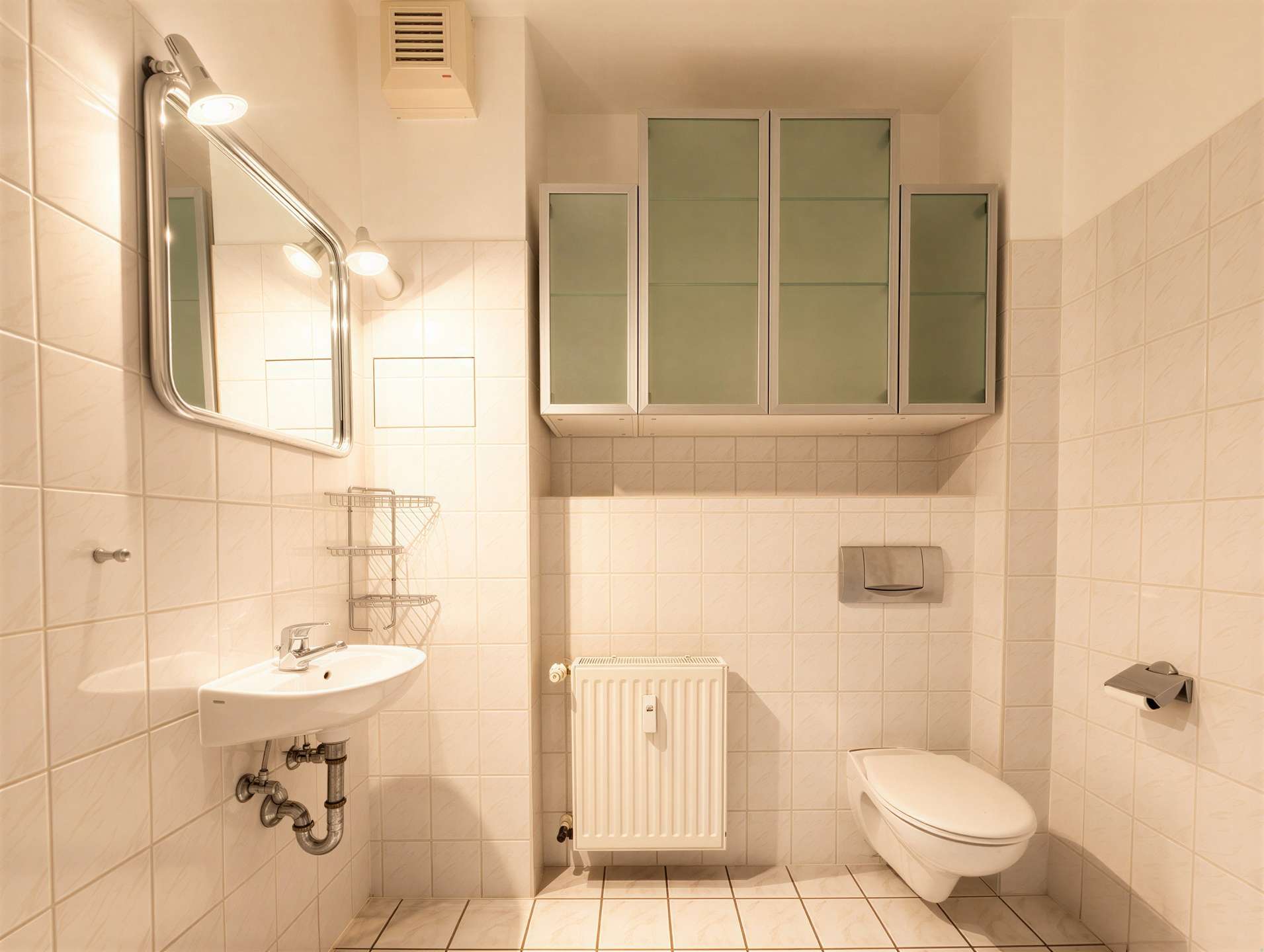 Bright white bathroom with a wall mirror, sink, metal storage shelf, radiator, and wall-mounted toilet.