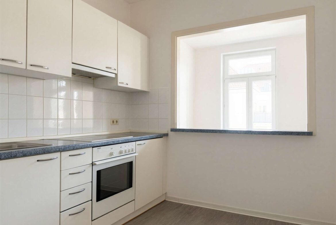 White kitchen with upper cabinets, a built-in oven and stove, speckled gray countertops, and a pass-through window to a bright adjacent room.