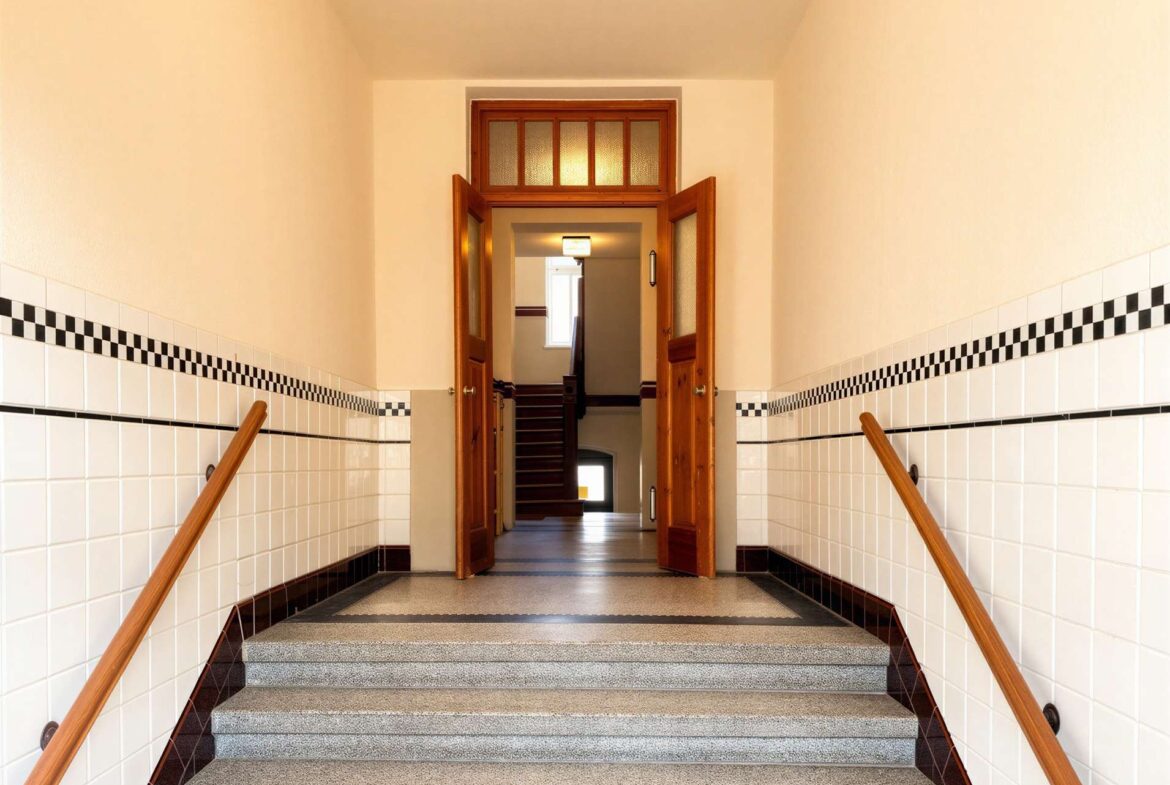 Interior stairwell with white tiled walls, wooden handrails, and open double doors at the top of a short flight, leading to more stairs down.