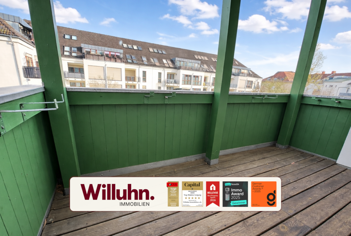 Balcony with green railing and wooden deck, overlooking apartment buildings under a blue sky; a real estate sign reads Willuhn Immobilien on the floor.
