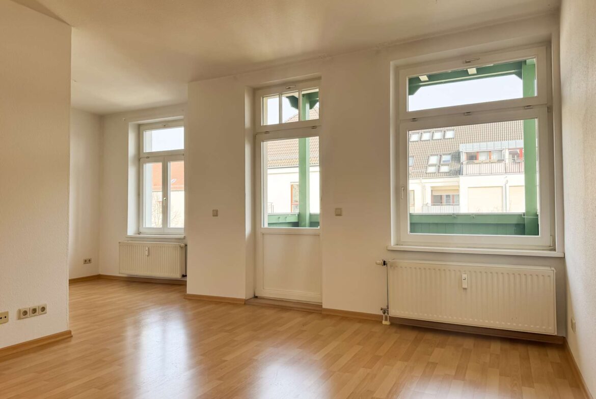 Bright empty room with large double windows letting in sunlight; light wood floor and white walls.