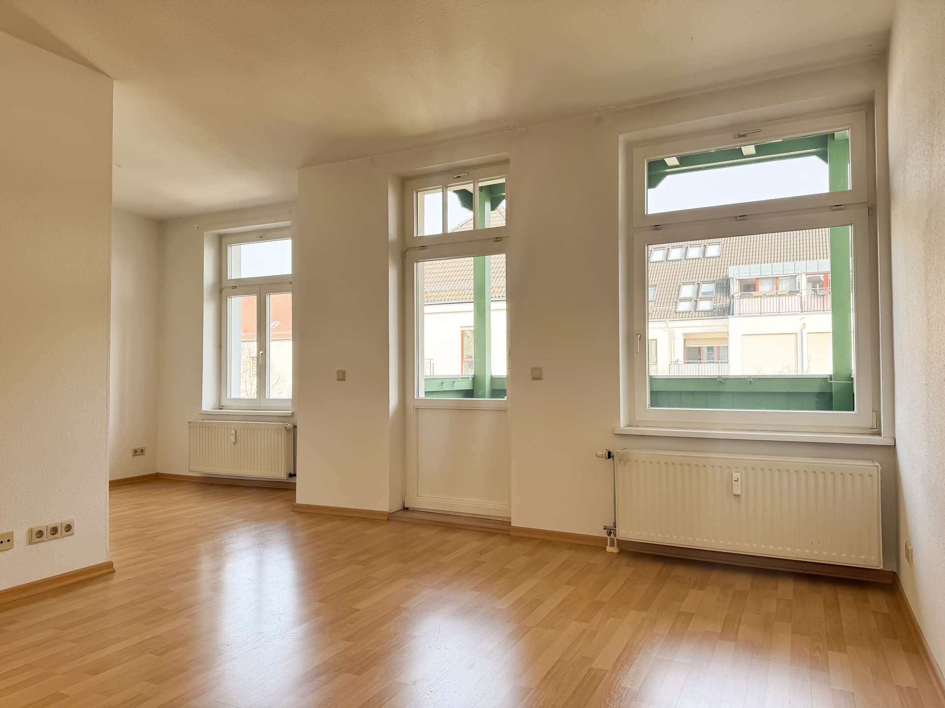 Bright empty room with large double windows letting in sunlight; light wood floor and white walls.