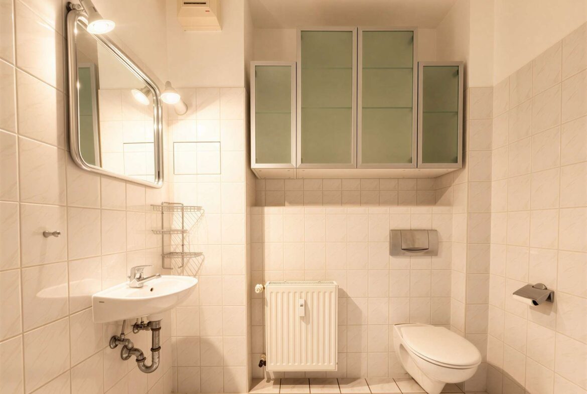 White-tiled bathroom with a wall-mounted sink, oval mirror, frosted glass wall cabinets, a toilet, and a radiator beneath a windowed shelf area.