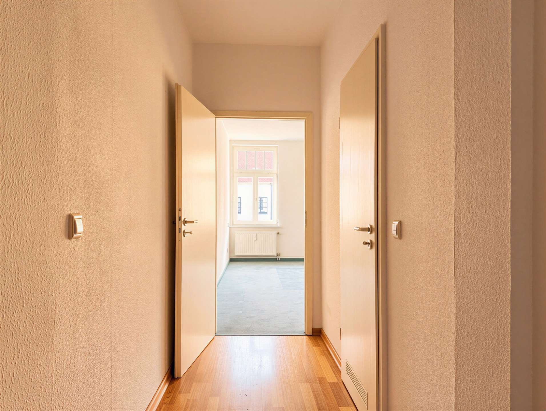 Narrow beige hallway with multiple open doors, leading to a sunlit room with a window in the distance.