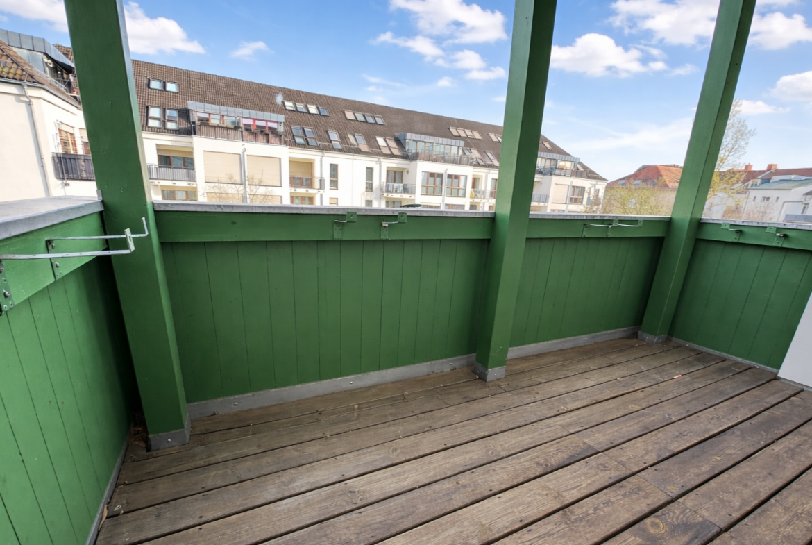 Green-painted wooden balcony with vertical boards and railing, overlooking neighboring apartment buildings under a bright blue sky with clouds.