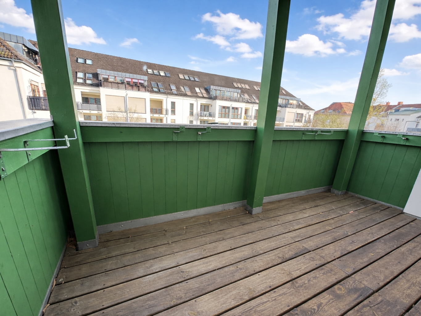 Green-painted wooden balcony with vertical boards and railing, overlooking neighboring apartment buildings under a bright blue sky with clouds.