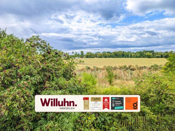 Rural field with a Willuhn Immobilien advertising sign in the foreground and cloudy sky above.