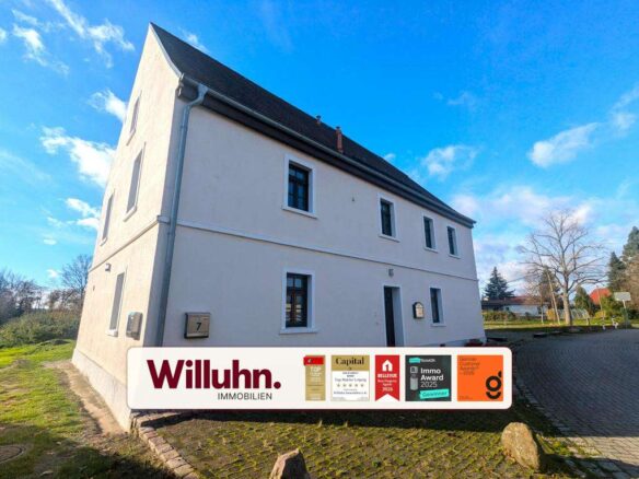 Exterior view of a white two-story house with a dark gabled roof under a blue sky; a Willuhn Immobilien banner covers the lower front.