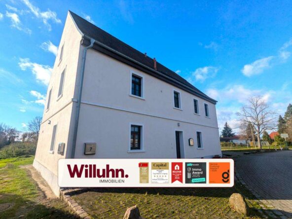 Exterior view of a white two-story house with a dark gabled roof under a blue sky; a Willuhn Immobilien banner covers the lower front.