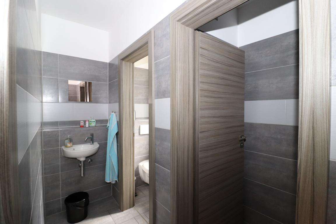 Small bathroom with gray tile walls, white wall-mounted sink, blue towel hanging, and a doorway to a toilet area visible through a partial wall.