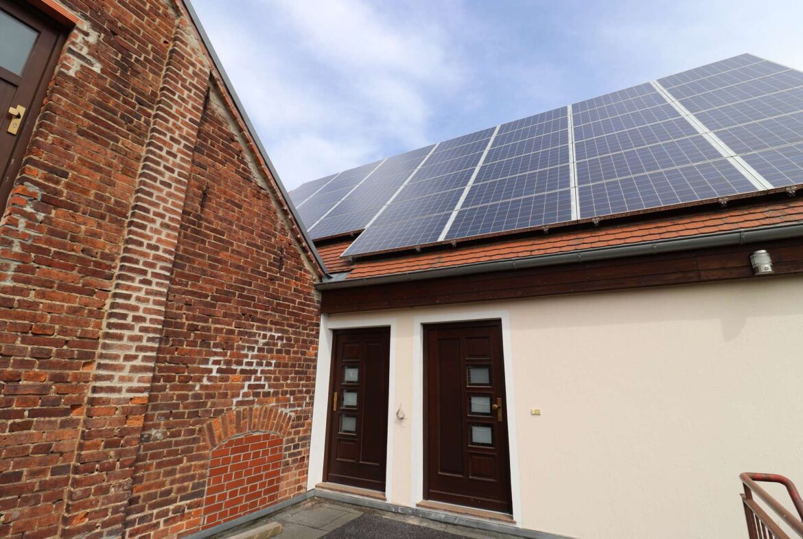 Brick building with a sloped roof covered in solar panels above two dark wooden doors; blue sky overhead.