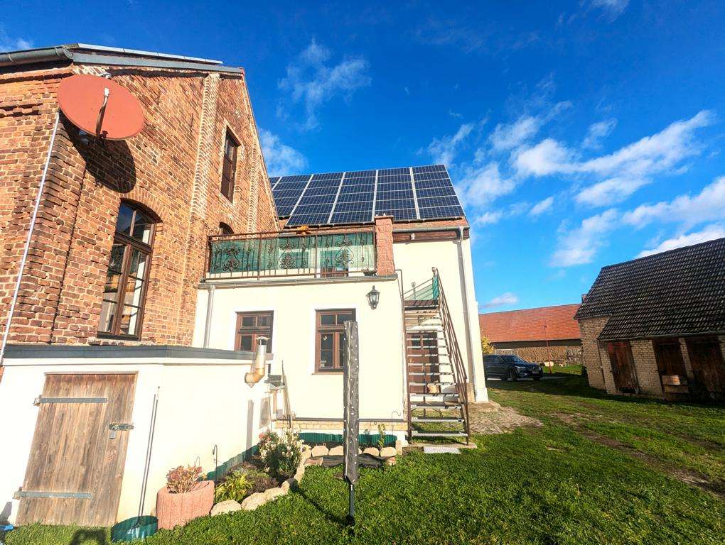 Brick house with solar panels on the roof, a red satellite dish, and an outdoor metal staircase beside a white extension under a blue sky.