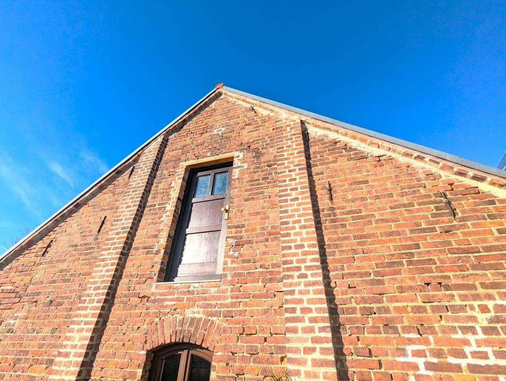 Brick building facade with a pitched roof, a wooden door on the upper level, and an arched doorway below under a clear blue sky.