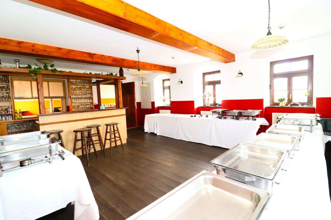 Buffet setup in a bright room with chafing dishes on long tables and a wooden bar with stools nearby.