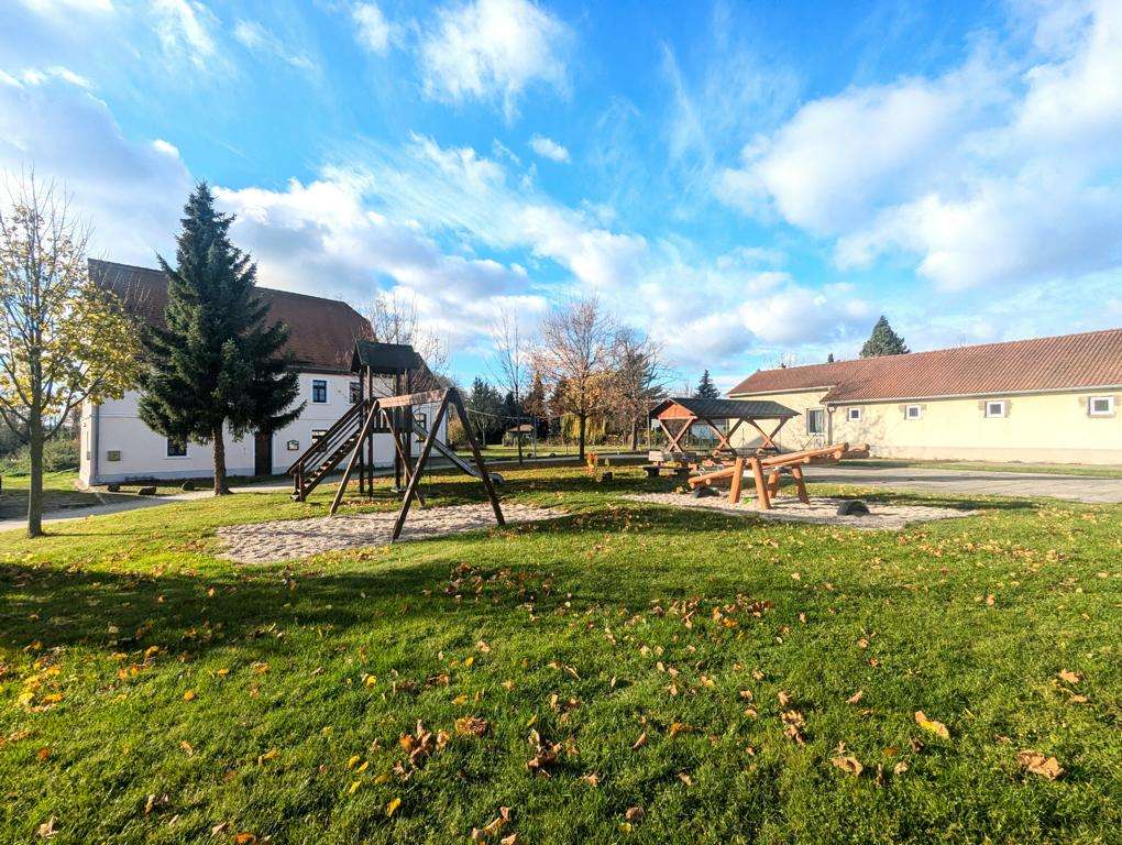 Sunny park with wooden playground equipment (swings and seesaw) on green grass near white buildings.
