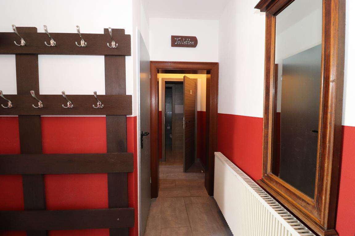 Hallway with a wooden coat rack on the left, red wainscoting, and an open door at the end labeled Toiletten above the frame.
