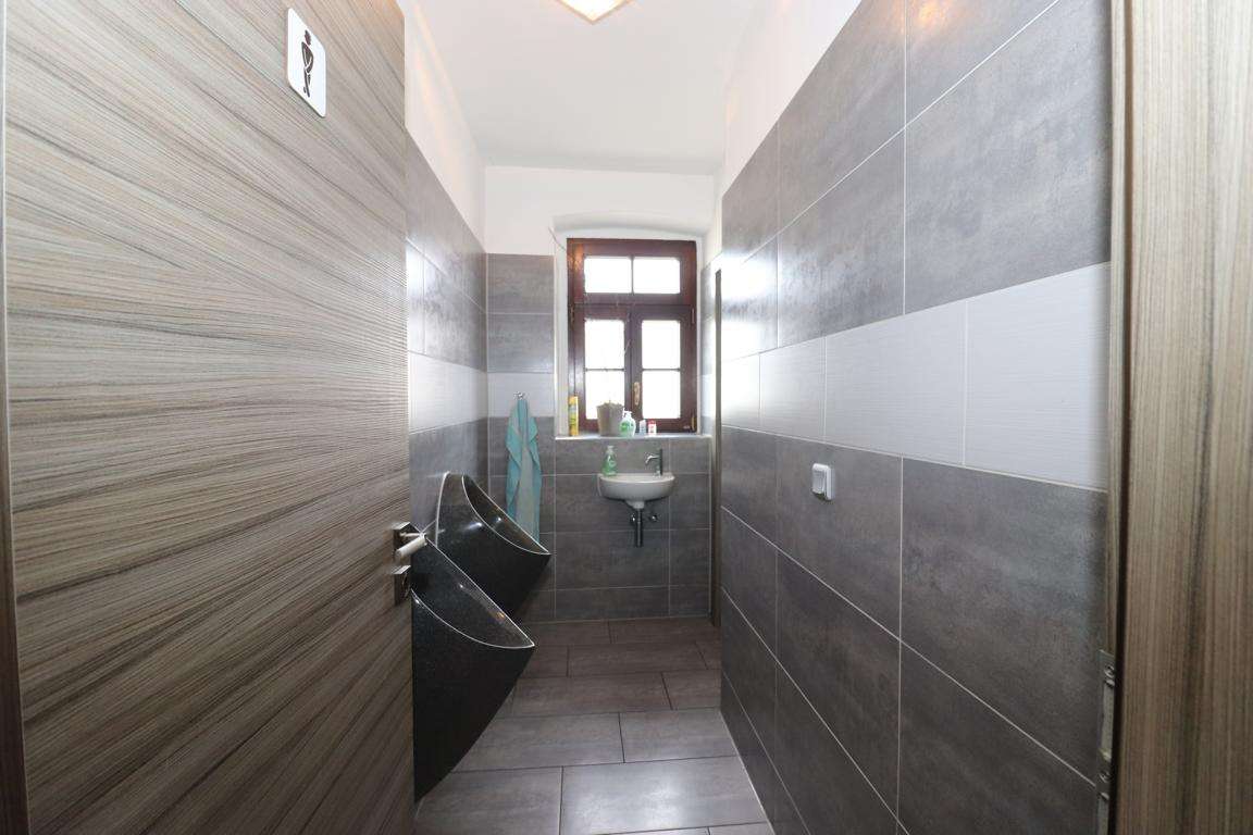 Narrow public restroom with two black urinals on the left, a small sink under a window at the far end, and gray tiled walls with white horizontal bands.