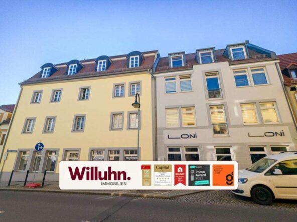 Three-story yellow and white buildings on a sunny street; a large Willuhn Immobilien banner covers the sidewalk.