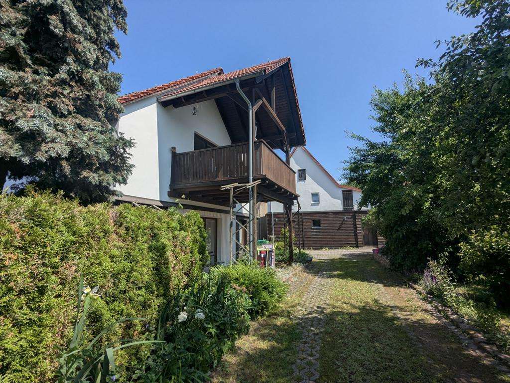 Two-story house with a wooden balcony and red tile roof, a stone path, and well-kept hedges on a sunny day.
