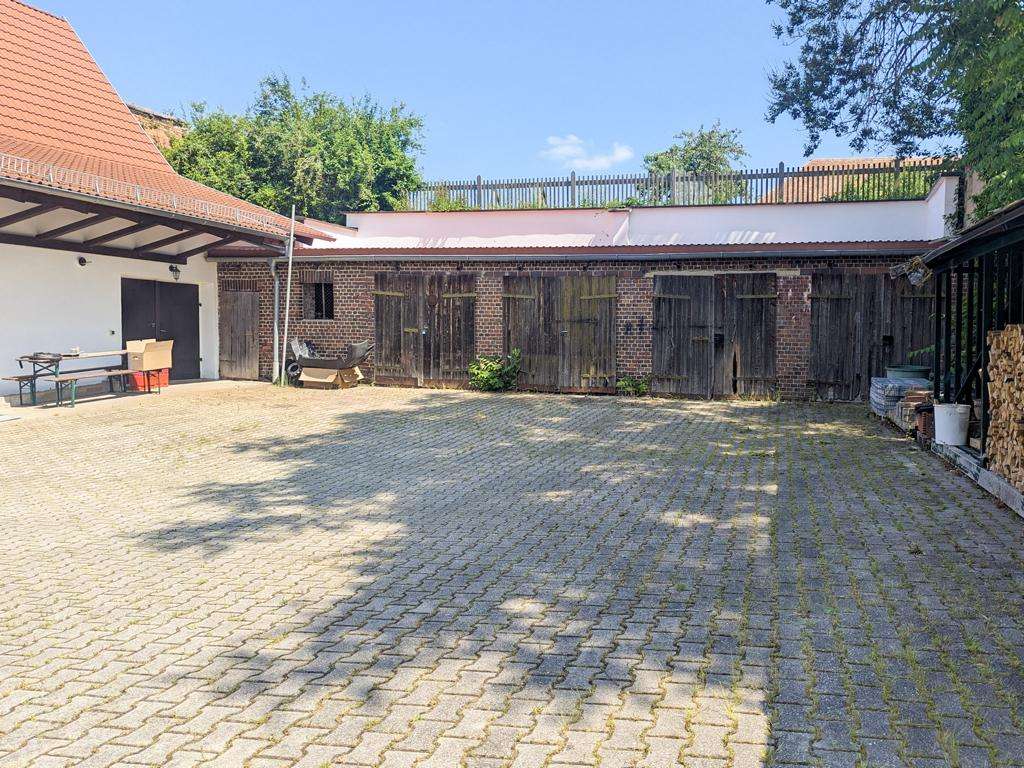 Sunny courtyard with a row of weathered wooden doors set into a brick building, a table and chairs on the left, and a woodpile on the right under a blue sky.