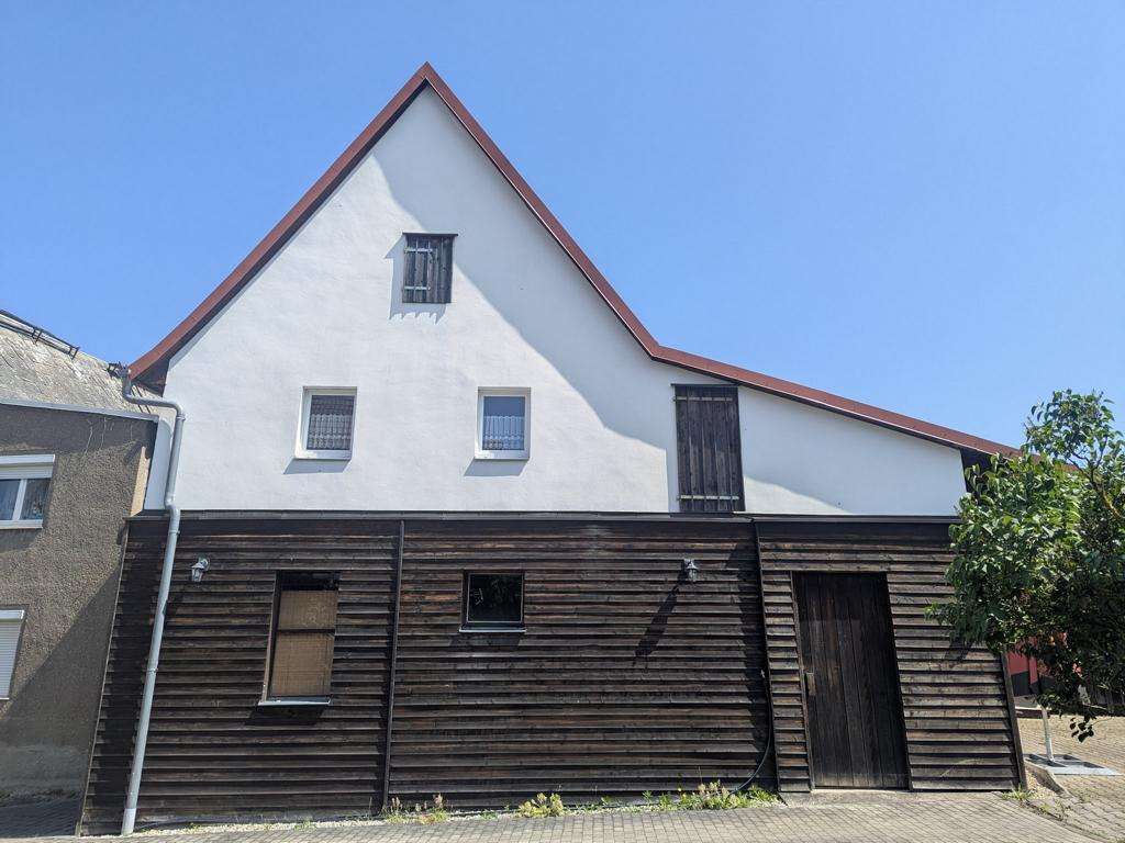 Two-story house with a white upper wall and dark wooden lower wall, a steep red-tiled roof, and barred windows against a blue sky.