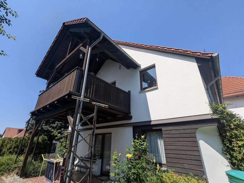 Two-story house with white and dark wood siding, elevated balcony on stilts and a clear blue sky above, in a sunny garden.