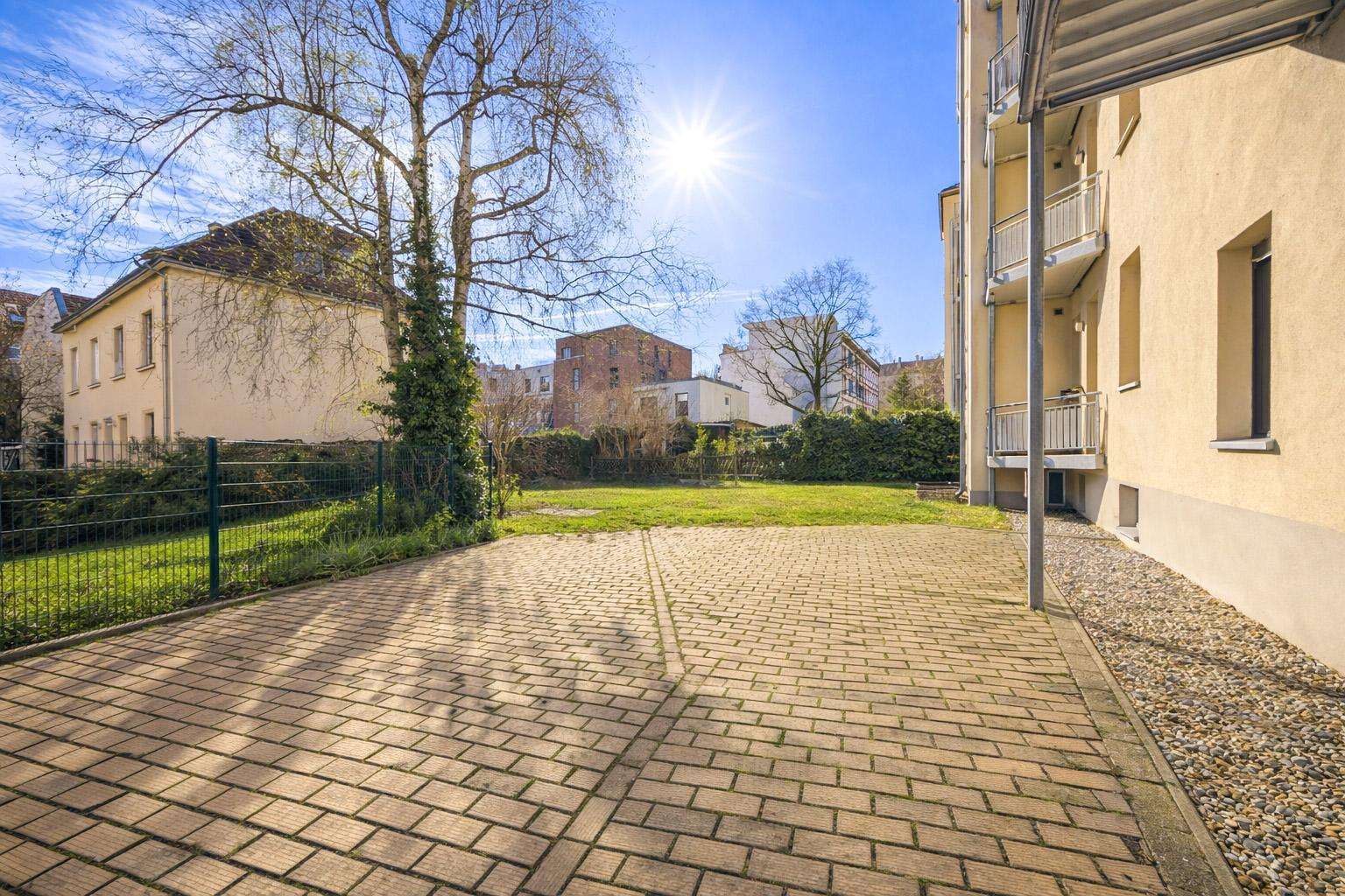 Sunlit residential courtyard with brick-paved path, beige building on the right, and bare trees over a green lawn in the background
