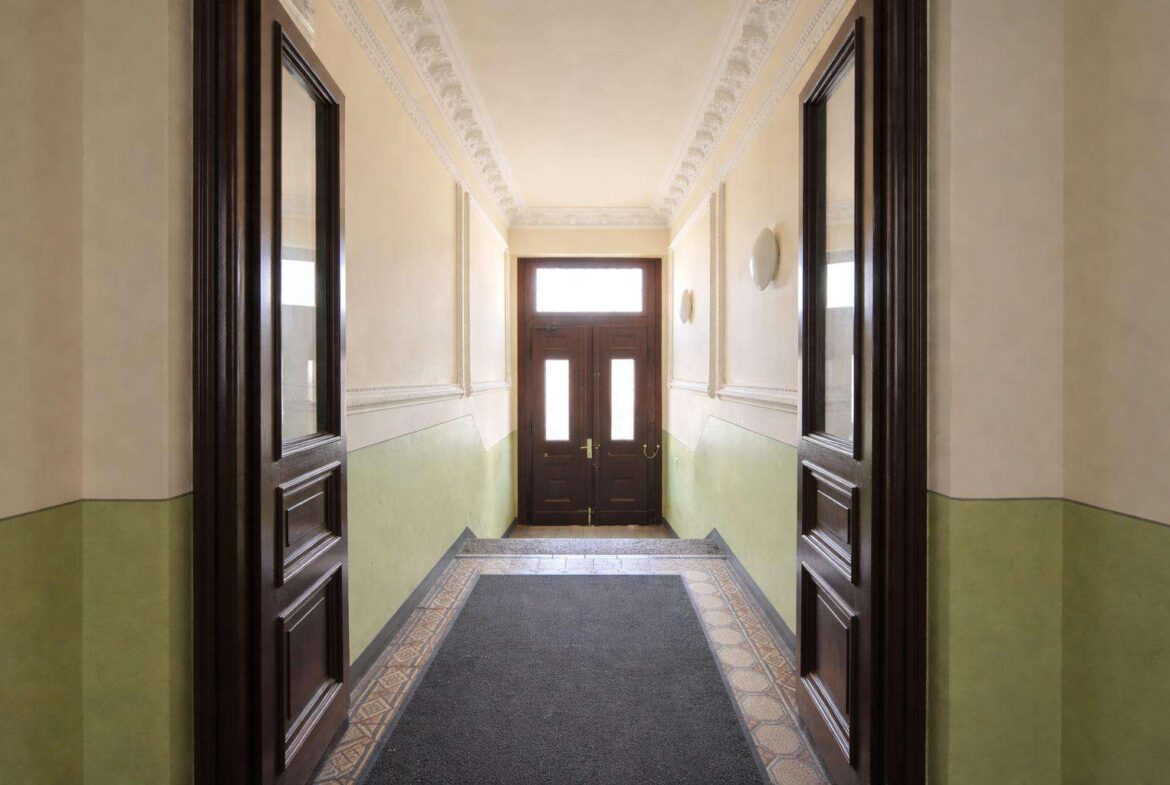 Narrow hallway in a historic building with decorative crown molding, cream walls, green wainscoting, and dark wood doors at the end.