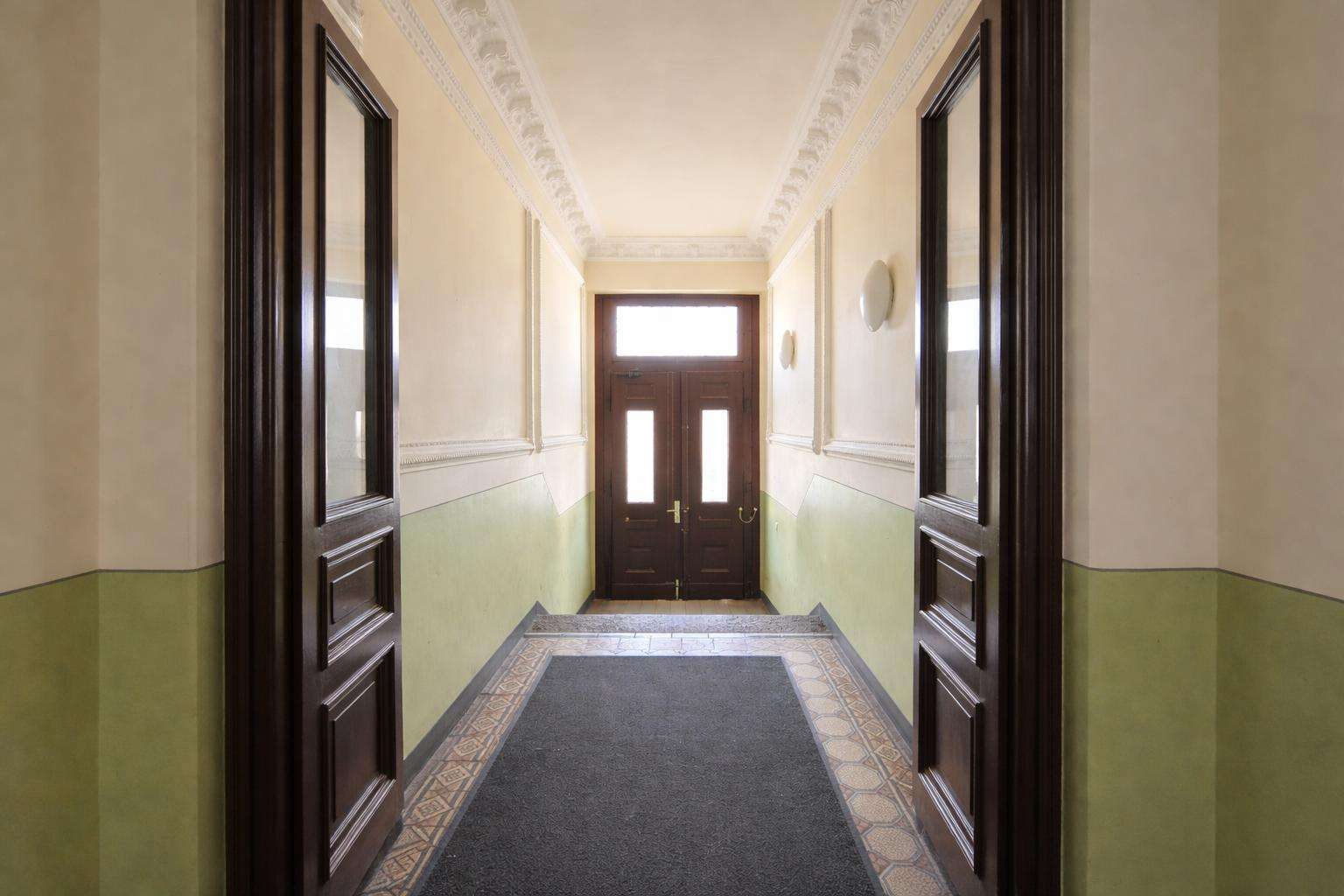 Narrow hallway in a historic building with decorative crown molding, cream walls, green wainscoting, and dark wood doors at the end.