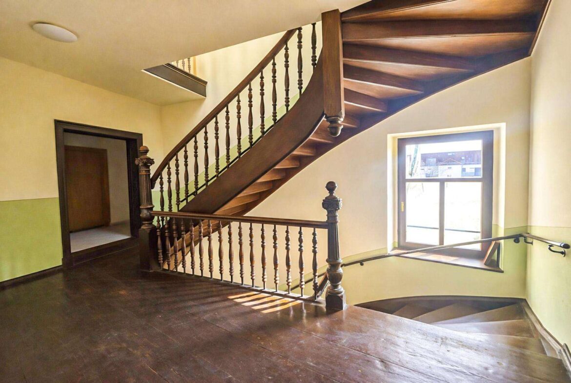 Curved wooden staircase with ornate balusters in a bright stairwell, sunlight from a nearby window.