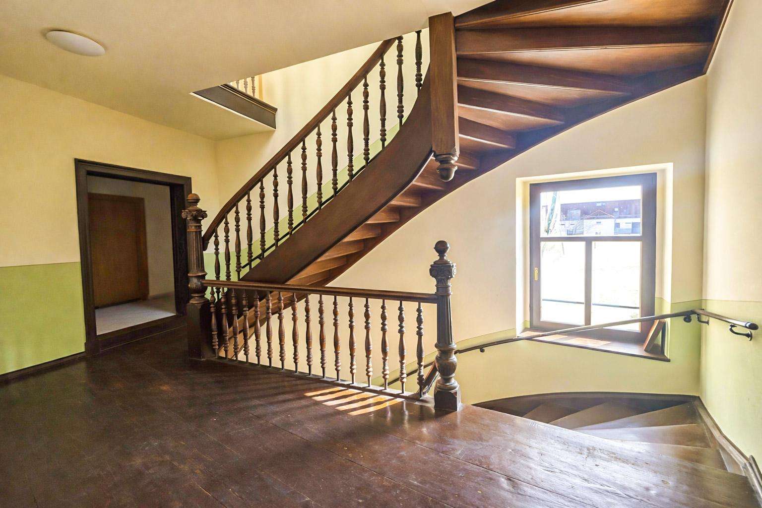 Curved wooden staircase with ornate balusters in a bright stairwell, sunlight from a nearby window.