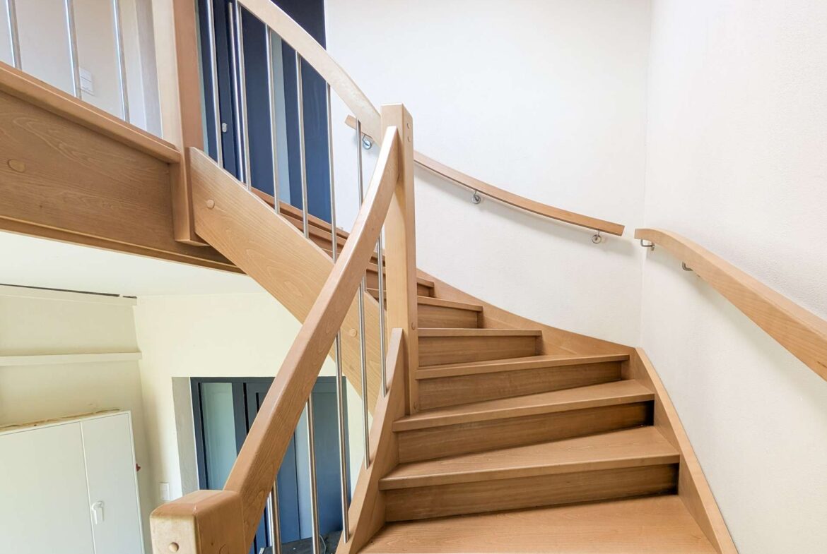 Indoor curved wooden staircase with metal balusters, leading to an upper floor in a bright interior. Downstairs blue doors are visible through the railing.