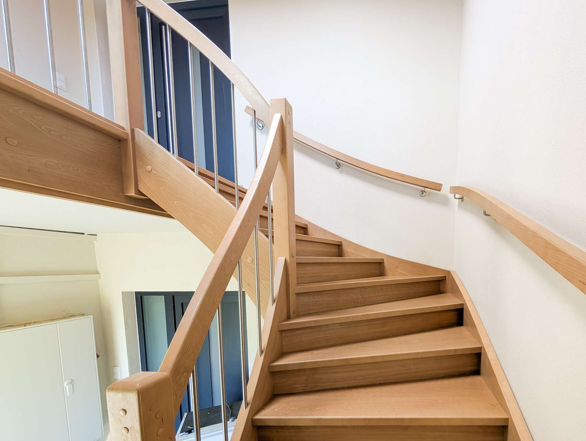 Indoor curved wooden staircase with metal balusters, leading to an upper floor in a bright interior. Downstairs blue doors are visible through the railing.