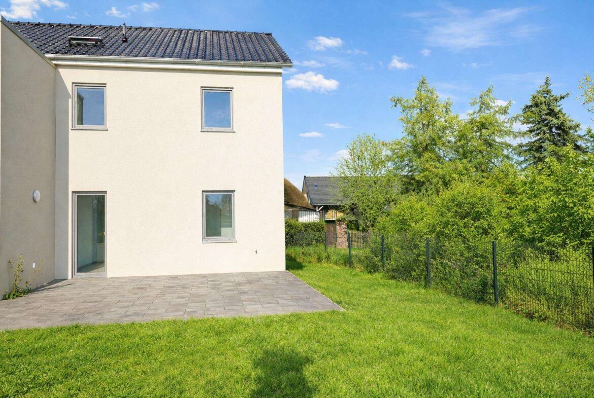 Backyard of a modern two-story house with a gray-tiled patio, beige exterior, green lawn, and a wire fence with trees beyond it.