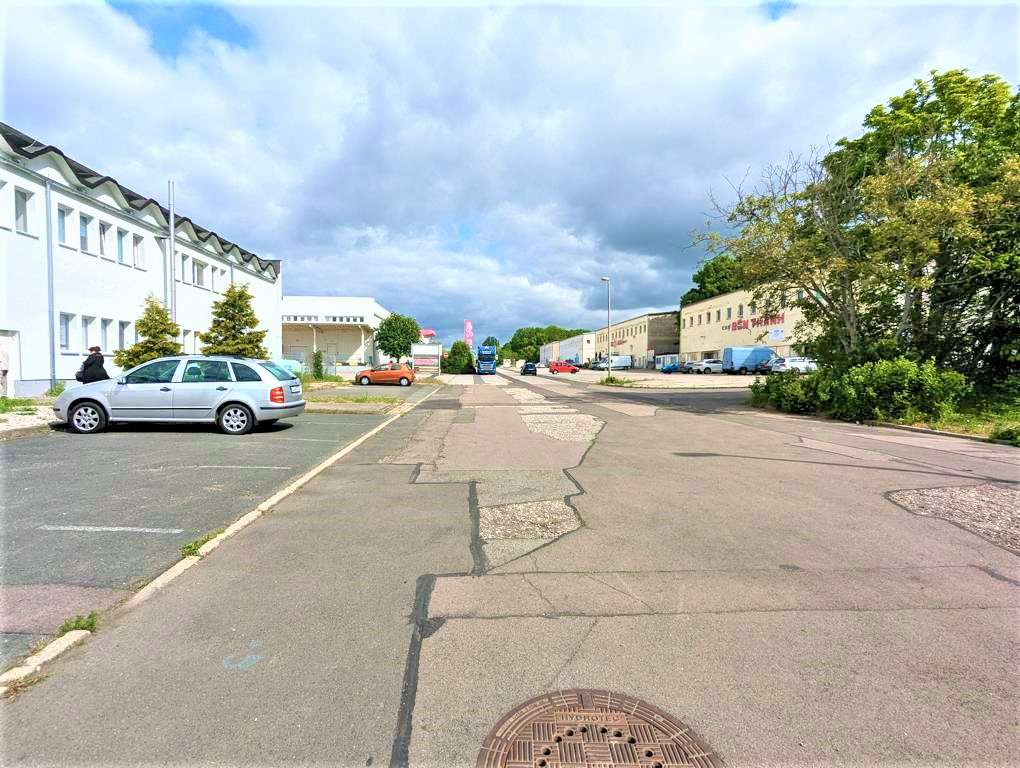 Wide street in an industrial area with white warehouse buildings, parked cars, and a cloudy sky.