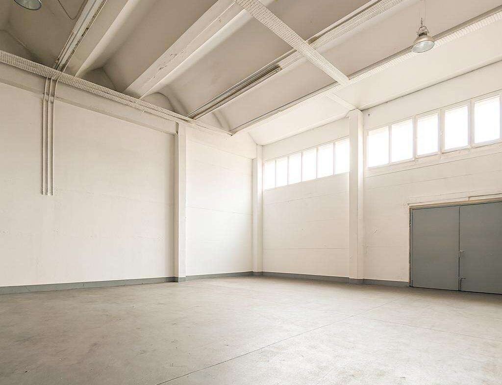 Empty industrial warehouse interior with a high ceiling, white walls, and a large gray double door on the right; row of windows along the upper wall and a clean concrete floor.