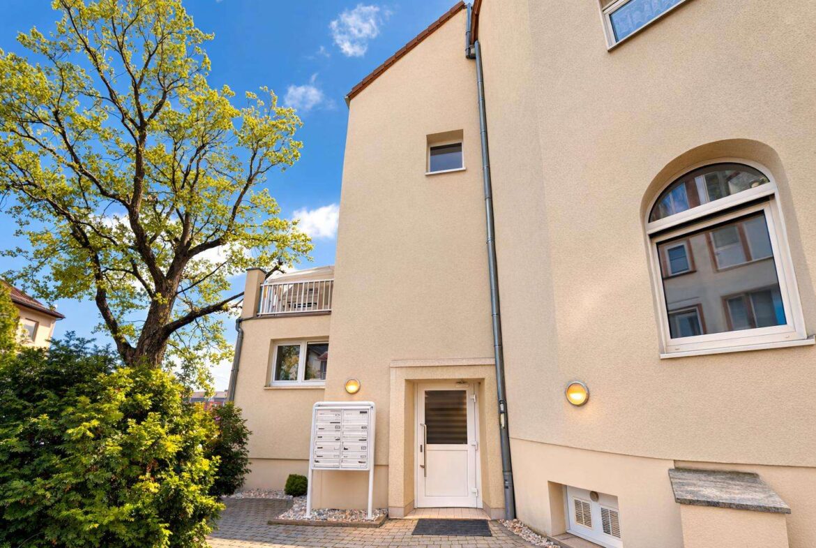 Beige apartment building entrance with white door, mailboxes, and a large tree to the left.