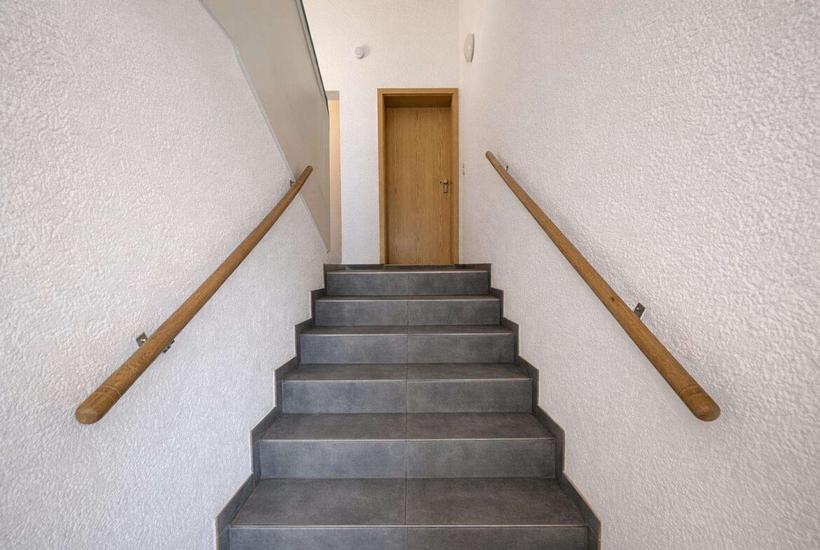 Stairwell with gray tiled steps, white textured walls, and wooden handrails on both sides leading to a wooden door at the top of the stairs.