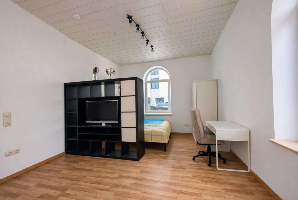 Small studio with a black modular shelving unit holding a TV, a bed by an arched window, and a white desk with a beige chair on wood floors.
