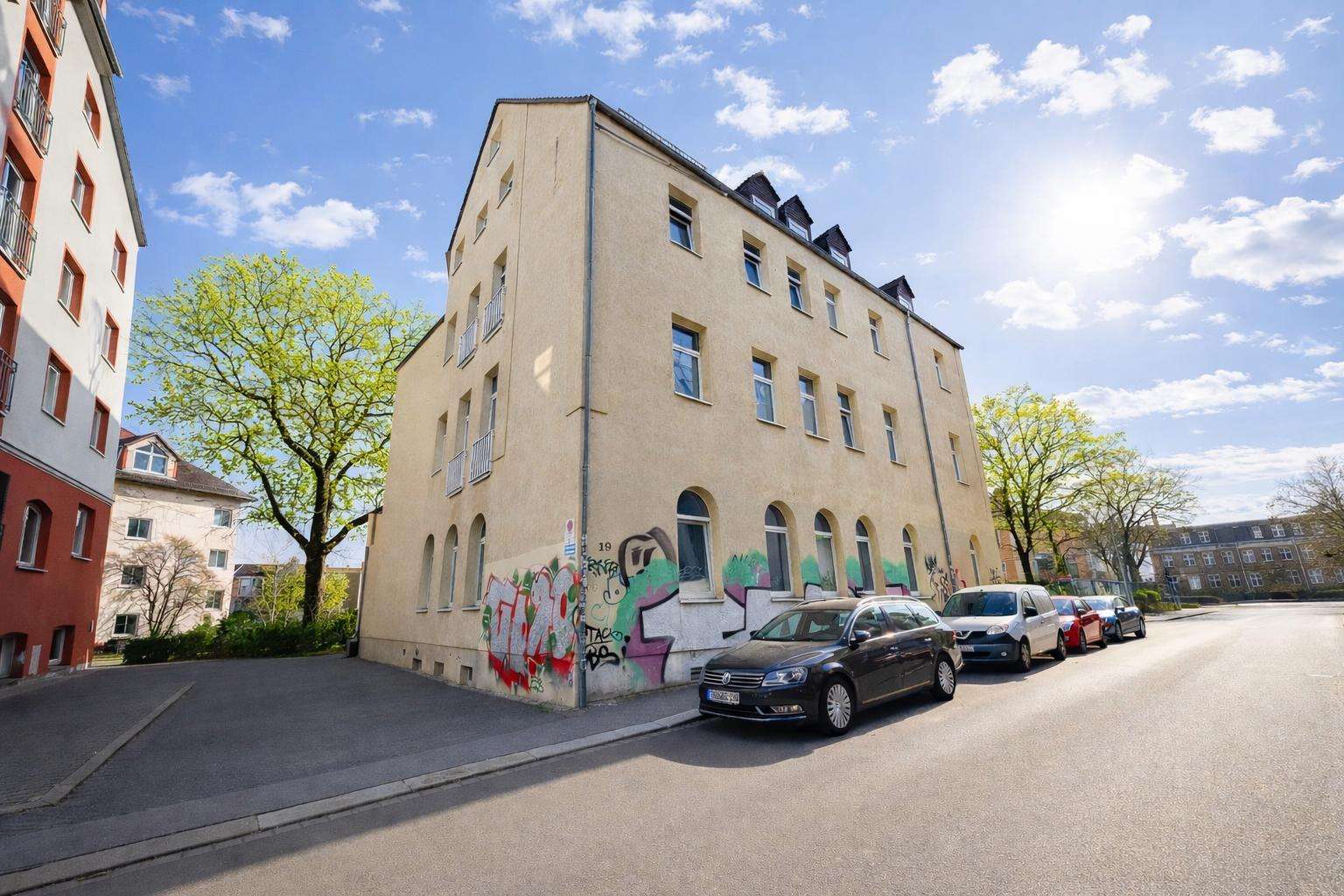 Three-story beige building with graffiti along the ground floor, parked cars lining the street, and a bright blue sky.
